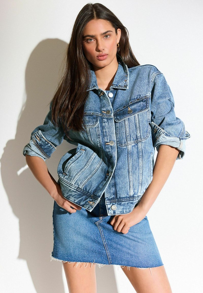 Chaqueta de mezclilla azul clara con un corte relajado, dos bolsillos en el pecho y puños deshilachados. Combinada con una falda de mezclilla azul clara a juego.