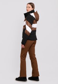 Gepolsterte Jacke in Schwarz, Weiß und Braun mit Zickzackmuster; verfügt über eine Kapuze und einen Reißverschluss. Kombiniert mit braunen Hosen und schwarzen Stiefeln.