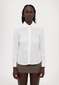 Hvit langermet bluse med klassisk krage, knapper foran og myk stoff. Kombinert med en khaki rutete miniskjørt.