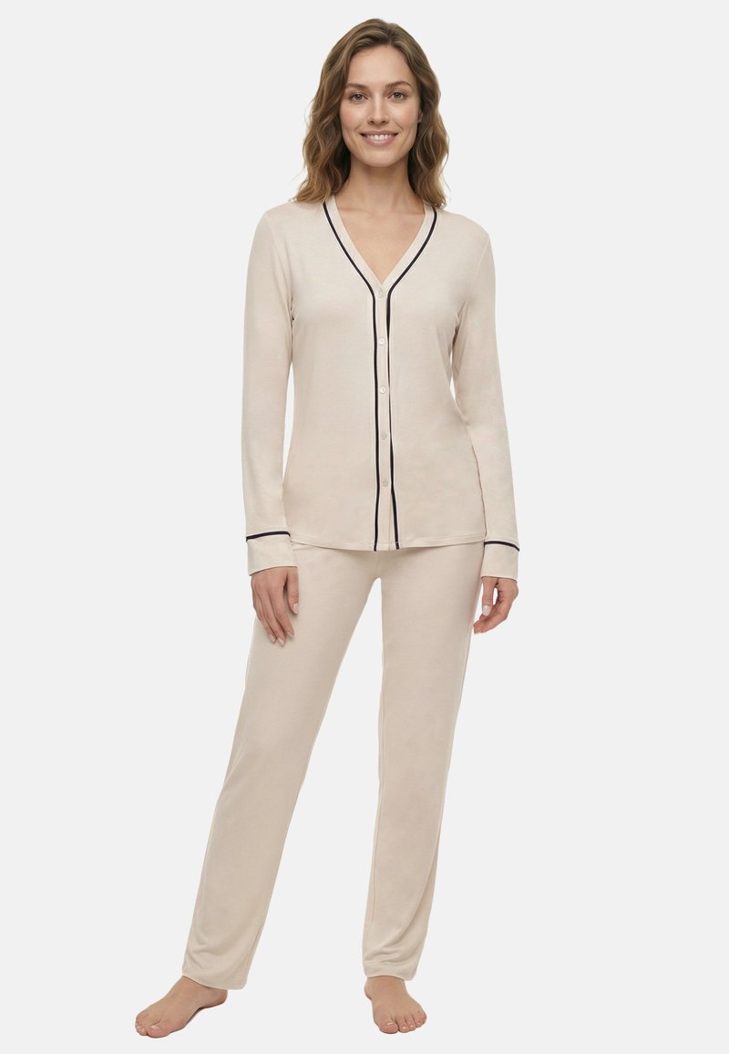 Nő áll mezítláb, bézs hosszú ujjú, gombos felsőt és hozzáillő nadrágot visel, fekete szegéllyel az ujjvégeken és az elején.