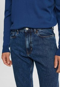 Persona vestida con pantalones vaqueros oscuros y una camisa azul de manga larga, con las manos relajadas a los lados, mostrando la cintura y la parte superior de las piernas.