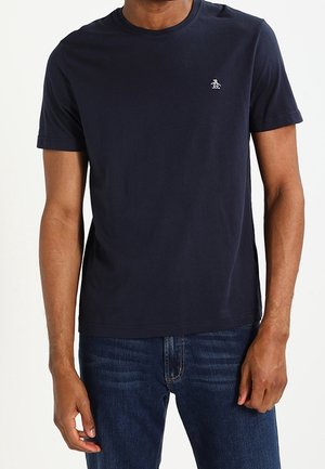 Homme portant un t-shirt uni bleu marine à manches courtes avec un petit logo blanc de pingouin sur la poitrine gauche et un jean bleu foncé.