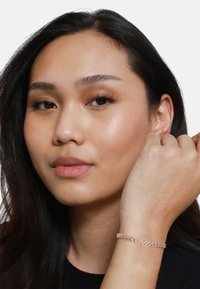 Bracciale in oro rosa con anelli intrecciati, indossato su un polso, presenta una texture liscia e una finitura lucida su una camicia nera.