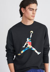 Μαύρο φούτερ με μακριά μανίκια που διαθέτει ένα πολυχρωμο λογότυπο Jordan. Ο σχεδιασμός περιλαμβάνει μια σιλουέτα σε ζωντανές αποχρώσεις στο σκοτεινό ύφασμα.