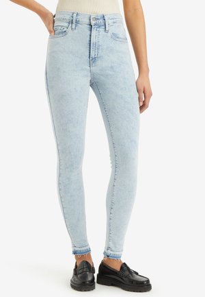 Femme portant un jean skinny taille haute bleu clair avec des ourlets effilochés et des mocassins noirs, debout avec une main dans la poche.