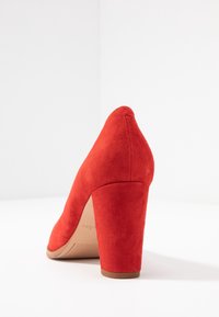 Chaussure à talon haut en daim rouge avec un bout pointu et un talon épais. La semelle est beige avec un léger relief antidérapant.