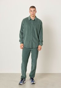 Zeleno korduroj srajco in ujemajoče hlače. Srajca ima gumbasto zasnovo s ovratnikom. Tekstura je rebrasta. Črni in modri superge.