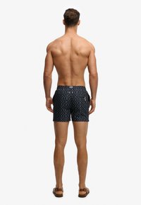 Mannequin masculin debout, dos visible, portant un short de bain noir à motifs et des sandales marron sur fond blanc.