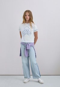 Weißes, kurzes T-Shirt mit besticktem Totenkopf- und Knochenmotiv; kombiniert mit hellblauen weit geschnittenen Jeans und einem lila Pullover, der um die Taille gebunden ist.