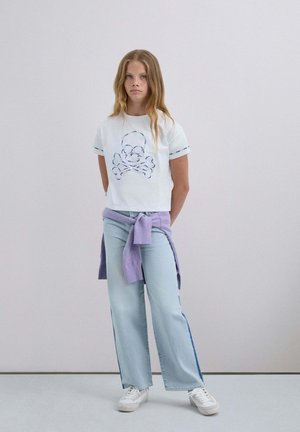 Camiseta blanca corta con un diseño bordado de calavera y huesos cruzados; combinada con unos jeans de pierna ancha de color azul claro y un suéter morado atado a la cintura.