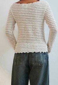 Off-white, gebreid sweater met een textuurpatroon en een gekartelde halslijn. Gecombineerd met hoog- taille, donkergrijze jeans met achterzakken.
