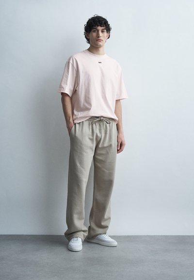 Νεαρός με σγουρά μαλλιά φοράει ανοιχτό ροζ φαρδιά μπλούζα, μπεζ παντελόνι με κορδόνι και λευκά αθλητικά παπούτσια, στέκεται μπροστά σε έναν απλό τοίχο.