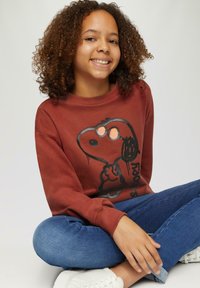 Bruine sweatshirt met een graphic van Snoopy; tegen een neutrale achtergrond. Draagt het met een blauwe jeans en witte sneakers, zittend met gekruiste benen.