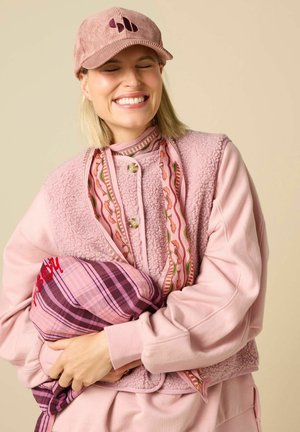 Femme souriante aux yeux fermés portant une casquette rose, une écharpe à motifs et un gilet texturé tout en serrant un coussin à carreaux violet avec une broderie rouge.