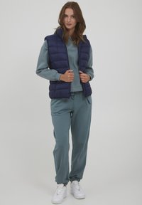 Navy puffer vest over een lichtteal sweatshirt en bijpassende joggers, met elastische boorden en tailleband, gecombineerd met witte sneakers.