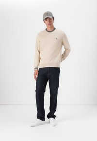 Bežový sveter s okrúhlym výstrihom, prúžkované manžety, v kombinácii s tmavomodrými džínsy a bielymi teniskami. Šedá čiapka dotvára outfit.