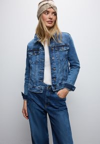 Giacca di jeans di un blu medio con chiusura a bottoni, tasche sul petto e cuciture a contrasto. Indossata sopra una maglietta bianca con jeans a vestibilità rilassata.