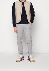 Chaleco sin mangas beige sobre una camiseta negra de manga larga, combinado con pantalones de chándal grises y zapatillas multicolores con detalles en rojo, azul y beige.