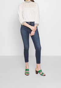 Light beige blouse with lace detailing and black trim, paired with dark blue skinny jeans and black, green, and white striped heeled sandals.