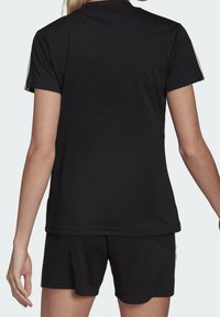 Kvinna i svart kortärmad polo skjorta och matchande svarta shorts, sedd bakifrån mot en enfärgad bakgrund.