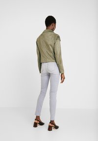 Veste en daim vert olive avec une silhouette ajustée, dotée d'accents texturés aux épaules. Associée à un jean slim gris clair et des sandales à talons noires.