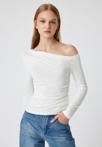 Witte langwerpige top met een off-the-shoulder ontwerp, voorzien van gerimpelde details aan de zijkant en gladde stof. Draagt met blauwe denim jeans.