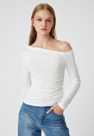 Witte langwerpige top met een off-the-shoulder ontwerp, voorzien van gerimpelde details aan de zijkant en gladde stof. Draagt met blauwe denim jeans.