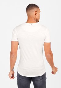 Camiseta blanca de manga corta con cuello redondo, diseño ajustado y textura sutil, combinada con jeans oscuros. Branding mínimo visible.