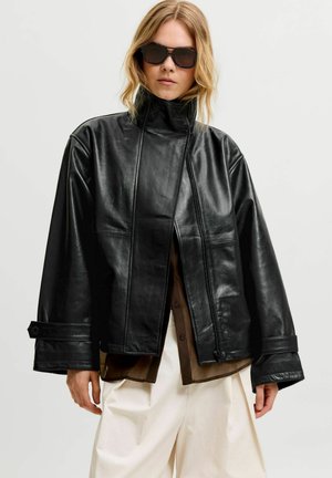 Donna bionda che indossa una giacca di pelle nera oversize, camicia marrone sotto, pantaloni bianchi e grandi occhiali da sole neri su sfondo semplice.