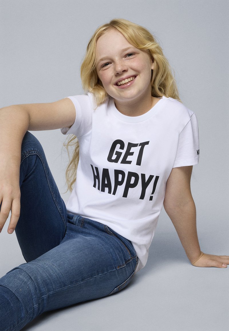 Vit t-shirt med kort ärm som har fet svart text "GET HAPPY!" på framsidan. Bärs tillsammans med blå jeans, i sittande pose mot en grå bakgrund.