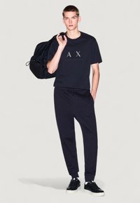 T-shirt noire avec le logo "AX", associé à un jogging noir et des baskets noires. La tenue comprend un sac à dos noir sur l'épaule gauche.