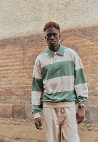 Jeune homme aux cheveux bouclés portant une chemise à manches longues rayée verte et crème ainsi que un pantalon beige, se tenant devant un mur en briques.