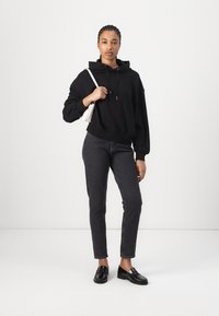 Zwarte hoodie met een subtiele logo, gecombineerd met donkere grijze slim-fit jeans en zwarte instapschoenen. Het model houdt een kleine witte handtas vast.