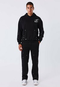 Zwarte hoodie met wit spetterontwerp, voorzak; gecombineerd met zwarte broek met vergelijkbaar spetterdetail. Model draagt witte sneakers.