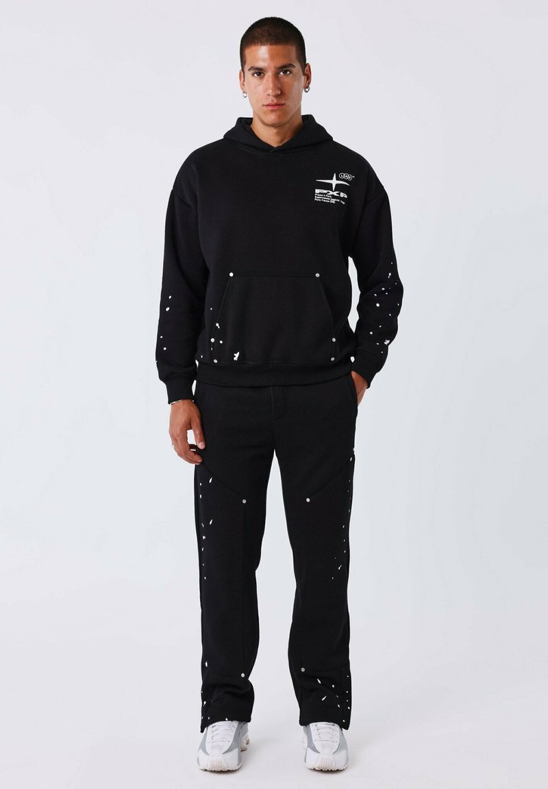 Schwarzer Hoodie mit weißen Farbspritzern, mit einer Vorderseite-Tasche und passenden schwarzen Hosen mit ähnlichen Mustern. Das Model trägt weiße Sneakers.