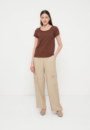 Mujer de pie con las manos en los bolsillos, lleva una camiseta marrón de manga corta, pantalones cargo beige de pierna ancha y zapatos beige tipo slip-on sobre fondo blanco.