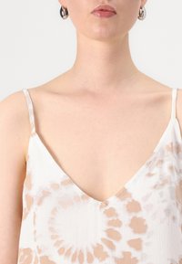 Valkoinen ja beige naruolkaiminen topi, jossa on syvä V-mallinen kaula-aukko, pehmeä ja teksturoidun tuntuinen kangas sekä hienovaraisia kuvioita.