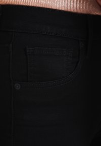 Pantalones vaqueros de mezclilla negra con un bolsillo frontal que presenta un sutil detalle de costura. Textura suave, diseño ajustado y un estilo clásico de cinco bolsillos.