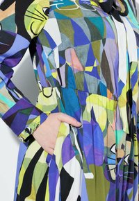 Abito colorato e decorato con forme geometriche in viola, verde, blu e nero. Maniche lunghe, vestibilità comoda, con tasche laterali e dettagli con bottoni.