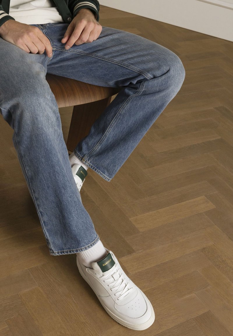 Baskets en cuir blanc avec des accents verts, portées avec un jean en denim bleu clair, posées sur un sol en bois chevron.