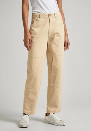 Pantaloni in cotone beige con vestibilità ampia, dotati di tasche laterali e dettagli cuciti. Abbinati a sneakers bianche.