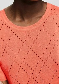 Gros plan d'une personne portant un haut en maille corail avec un petit motif ajouré en forme de losange et un col rond.
