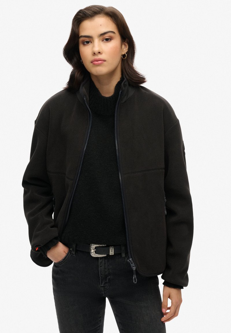 Giacca in pile nera con colletto alto, chiusura lampo frontale e tasche laterali. Indossata sopra un maglione nero a trama e abbinata a jeans neri.