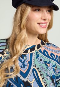 Haut à motifs colorés avec des dessins floraux et géométriques complexes en bleu, vert, rose et noir ; le mannequin porte une casquette noire.