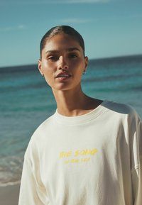 Prevelik pulover kremaste barve z rumenim napisom "PESEM MORJA." Gladek tekst, okrogli izrez, na ozadju plaže.