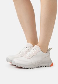 Ljusgrå sneakers med en strukturerad yta, med vita snören, en vadderad krage och en orange sula för kontrast.