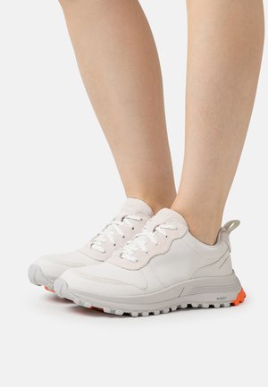 Lys grå sneakers med en tekstureret overflade, der har hvide snørebånd, en polstret krave og en orange ydersål for kontrast.