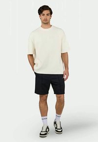 T-shirt texturizado de manga curta em creme, combinado com calções escuros e ténis preto e branco, de pé contra um fundo neutro.