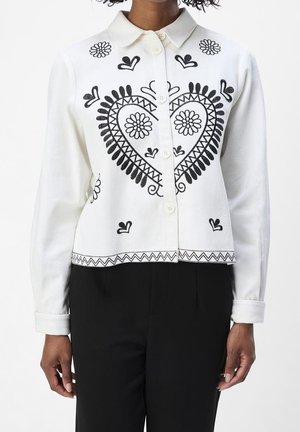 Cardigan blanc à boutons avec un cœur brodé noir et des motifs floraux, porté avec un pantalon noir.