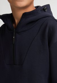 Felpa con cappuccio di colore blu navy, con chiusura a zip, caratterizzata da un tessuto testurizzato, dettagli delle cuciture e un cordino regolabile sul cappuccio.